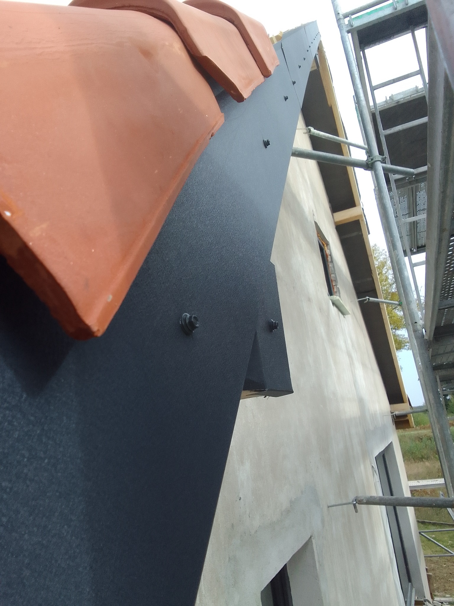 Montaż obróbki blacharskiej na dachu z częściowo ułożoną dachówką ceramiczną, widoczne rusztowanie i okno w stanie surowym.