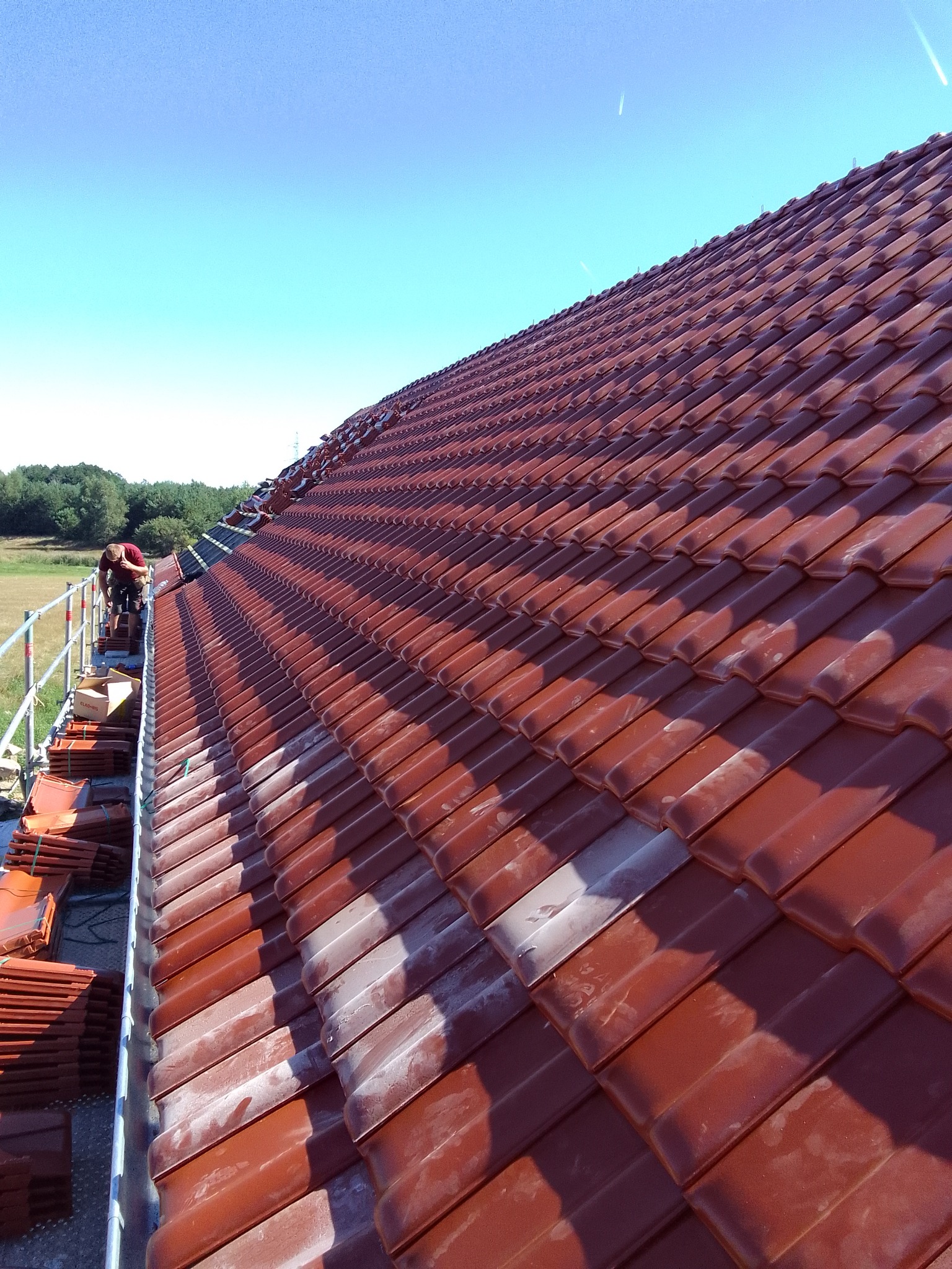 Czerwona dachówka ceramiczna układana na dachu, widoczny dekarz pracujący z rusztowania, błękitne niebo w tle.