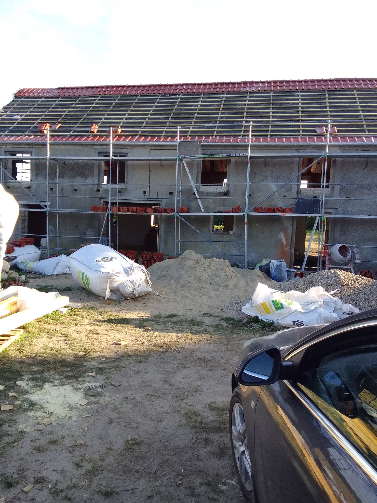 Budynek w trakcie prac dekarskich: położona więźba dachowa, częściowo ułożona czerwona dachówka, rusztowania, worki z materiałami budowlanymi i betoniarka na posesji, fragment samochodu...