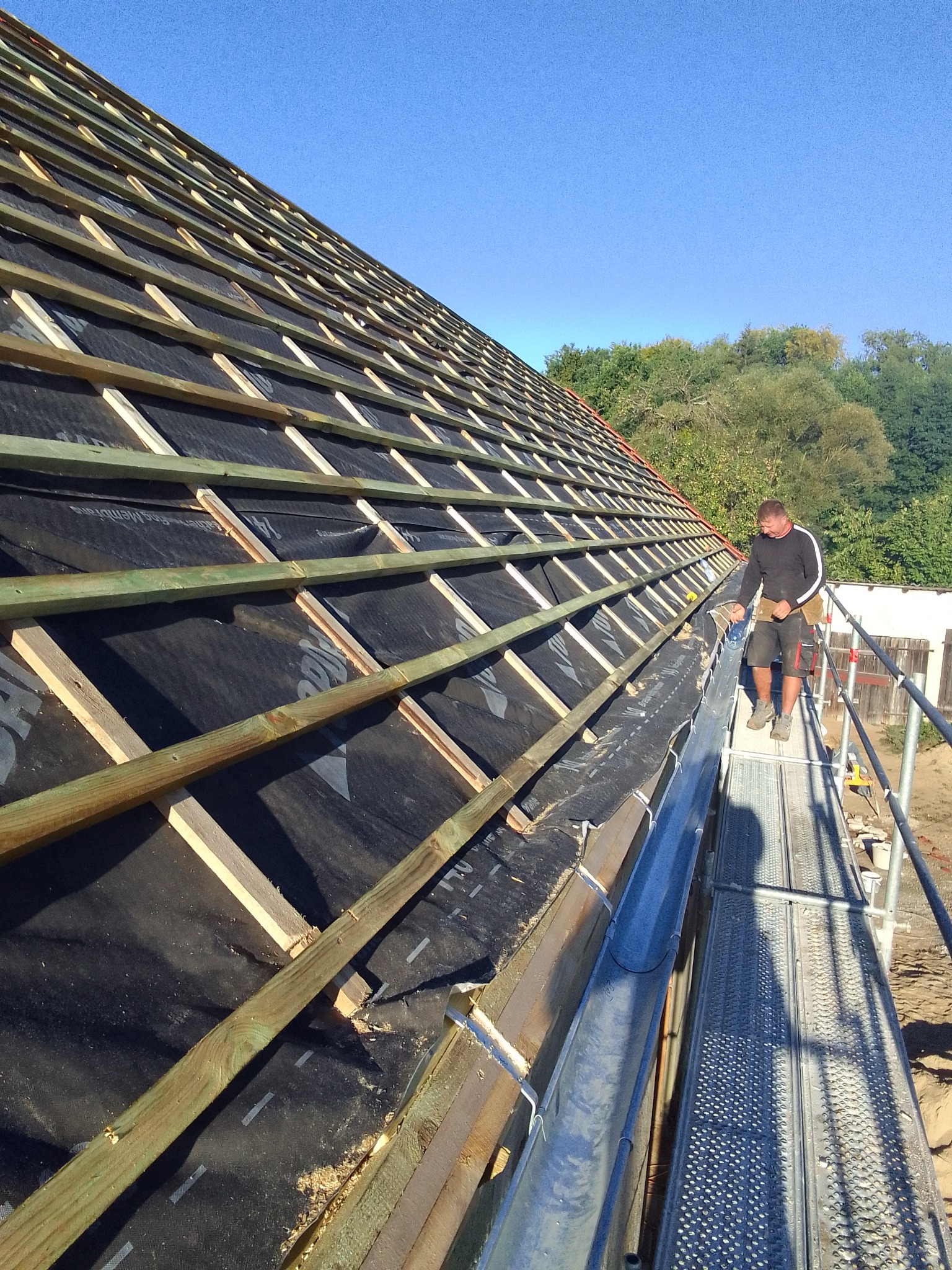 Montaż łat i kontrłat na membranie dachowej, widoczne rynny i pracownik na rusztowaniu w słoneczny dzień.