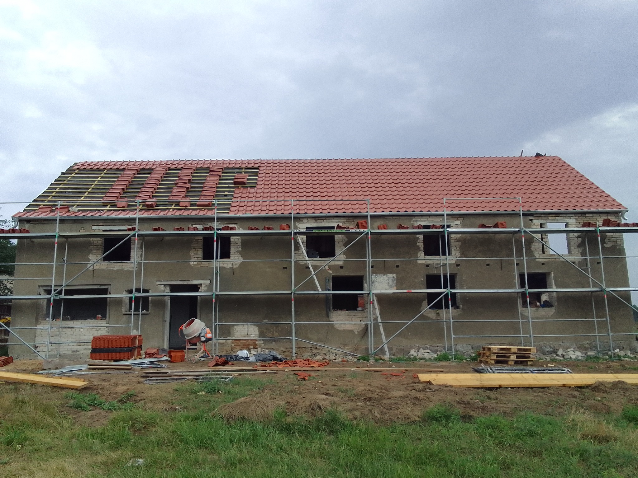 Dom w trakcie renowacji dachu, częściowo pokryty czerwoną dachówką, rusztowania na elewacji, widoczna betoniarka i paleta z dachówkami.