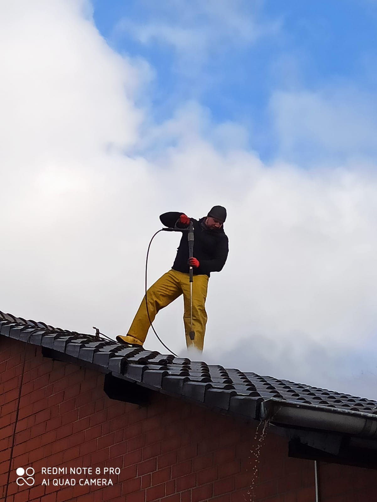 Pracownik w żółtych spodniach i czarnej kurtce czyści dach z czarnej dachówki za pomocą myjki ciśnieniowej na tle pochmurnego nieba.