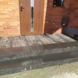 Nowe, dwustopniowe schody wejściowe z szarej kostki granitowej prowadzące do drewnianego podestu i brązowych drzwi wejściowych, na tle ceglanej ściany budynku.