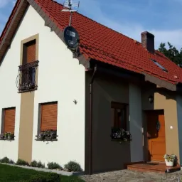 Dwukondygnacyjny dom z jasną elewacją, brązowymi akcentami i czerwoną dachówką, z oknami z roletami i balkonem z ozdobną balustradą, otoczony zielonym trawnikiem i roślinnością.