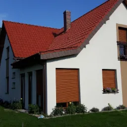 Elewacja domu jednorodzinnego w kolorze białym z brązowymi roletami, czerwoną dachówką i balkonem z kwiatami, widok z ogrodu, w tle drzewa.