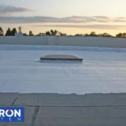 Biała, płaska powierzchnia dachu z świetlikiem, po renowacji, z widocznym logo firmy 'Hydron System' w dolnej części kadru, na tle zachodzącego słońca.