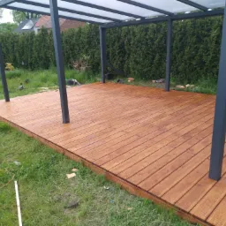 Świeżo zaimpregnowany taras drewniany z zadaszeniem w ogrodzie, widoczna butelka wody i radio budowlane.