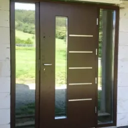 Brązowe drzwi wejściowe z przeszkleniami bocznymi, zamontowane w surowym stanie budynku, widok na zielony krajobraz przez szyby.