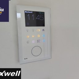 ELDAM Damian Szlendak - Panel sterowania Nexwell na ścianie, z wyświetlaczem czasu i ikonami funkcji inteligentnego domu. Biała obudowa, minimalistyczny design, nowoczesny interfejs.