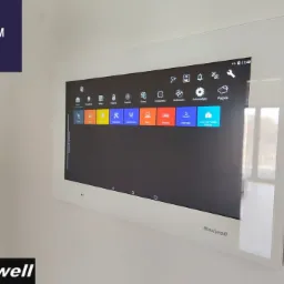 Biały panel dotykowy LCD 15,6 cala firmy Nexwell, zamontowany na ścianie, wyświetlający interfejs systemu smart home z ikonami różnych funkcji.