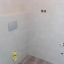 Wnętrze łazienki w trakcie remontu: ułożone jasne płytki imitujące kamień na ścianach, drewnopodobne panele na podłodze, widoczne żółte rury i przycisk spłuczki w zabudowie.