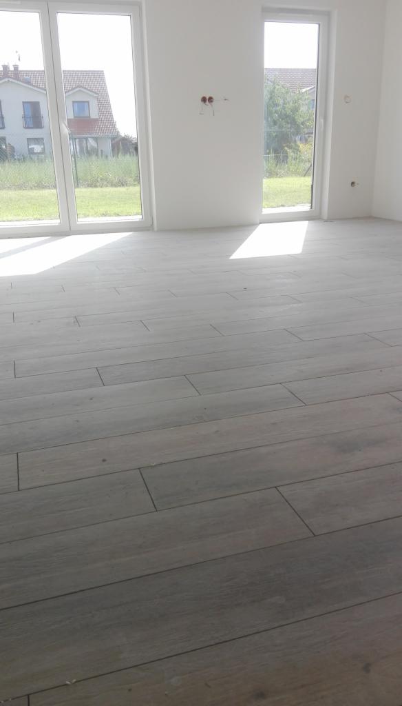 Jasne, podłużne płytki podłogowe imitujące drewno w nowym, pustym pokoju z dużymi oknami i widokiem na dom i trawnik na zewnątrz.