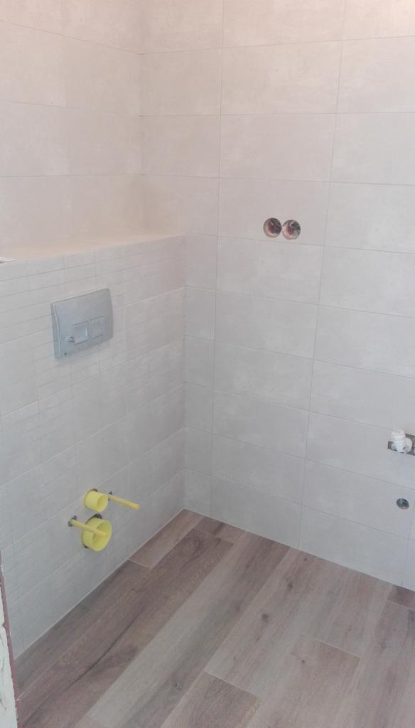 Wnętrze łazienki w trakcie remontu: ułożone jasne płytki imitujące kamień na ścianach, drewnopodobne panele na podłodze, widoczne żółte rury i przycisk spłuczki w zabudowie.