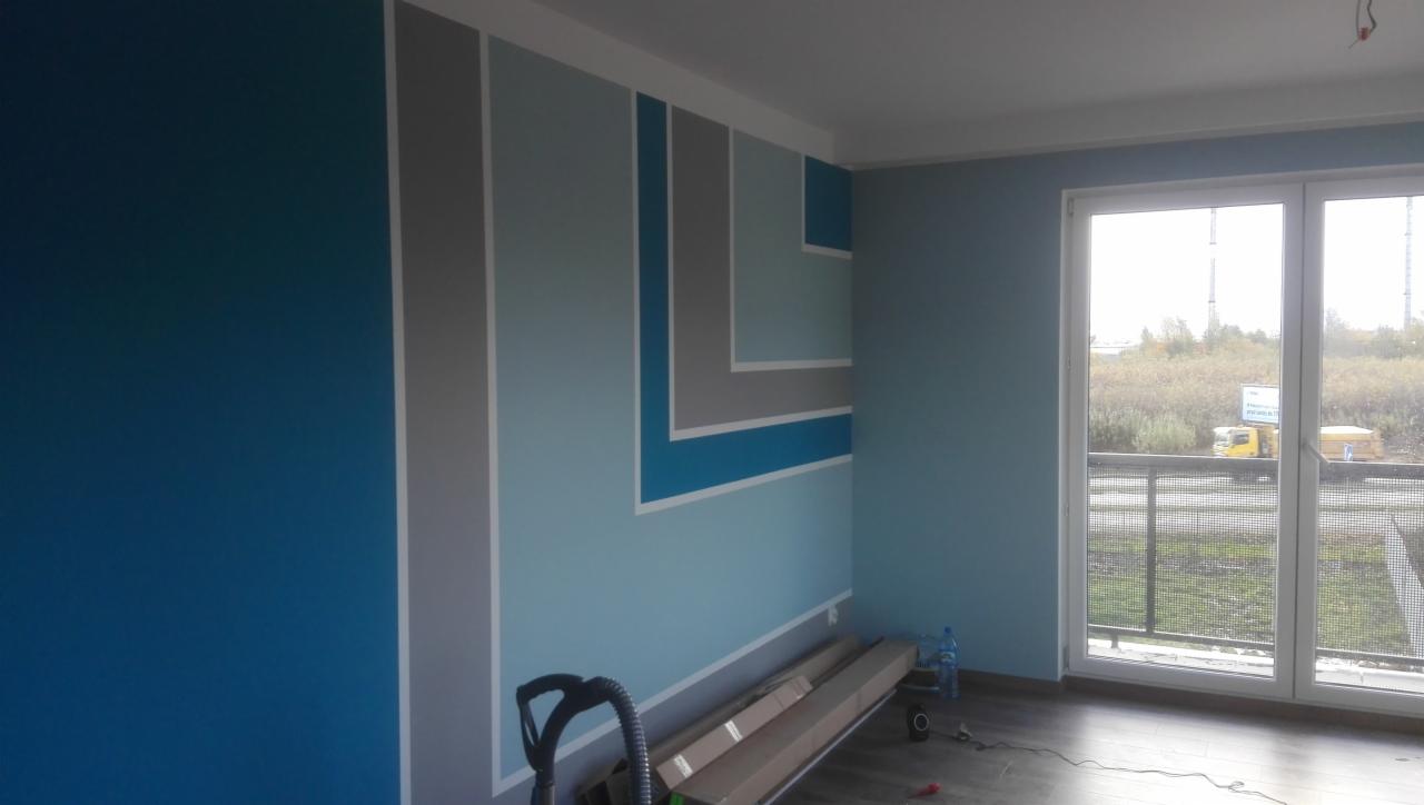 Wnętrze pokoju z pomalowanymi ścianami w odcieniach niebieskiego i szarego, z geometrycznym wzorem przy suficie. Widoczny odkurzacz i kartony na podłodze, okno balkonowe z widokiem na zewnątrz.