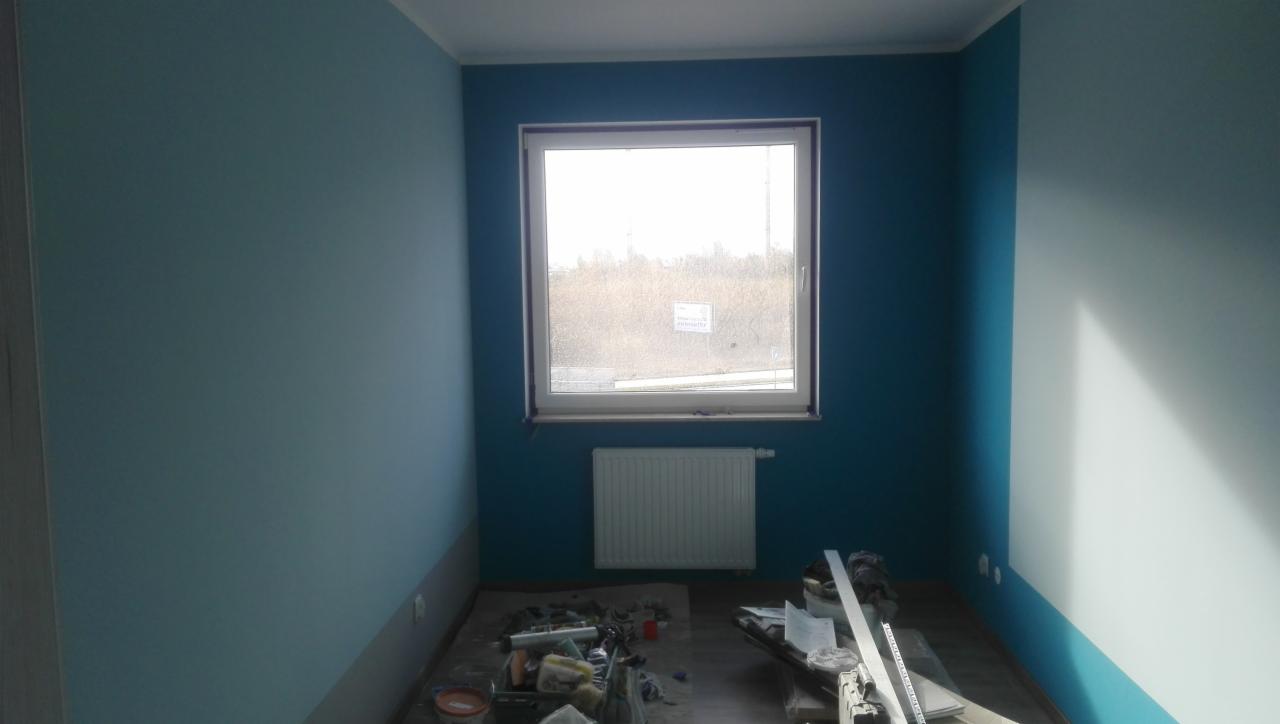 Pokój po częściowym pomalowaniu ścian na dwa odcienie niebieskiego, widoczne narzędzia malarskie na podłodze, okno z widokiem na zewnątrz.