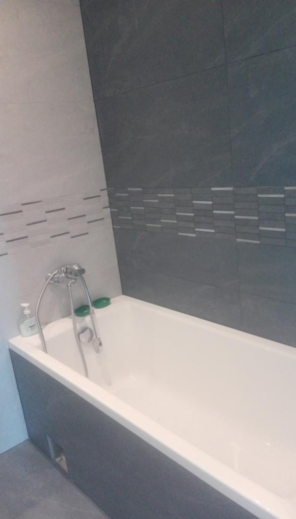 Wanna obudowana szarą płytą z baterią i mydłem, wykończona płytkami w dwóch odcieniach szarości z poziomym paskiem dekoracyjnym.