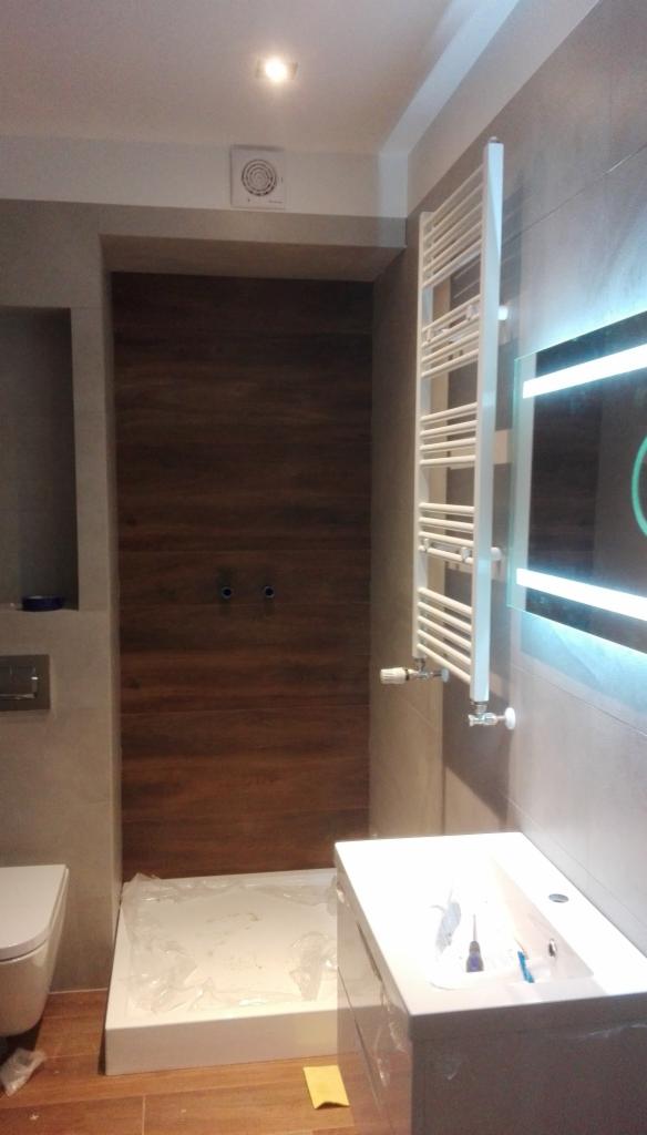 Montaż kabiny prysznicowej z drewnianą ścianą w łazience w Lubczynie, widoczny grzejnik drabinkowy i umywalka z podświetlanym lustrem, etap prac wykończeniowych.