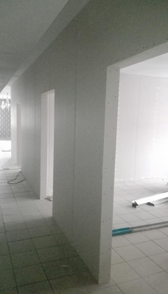 Korytarz w trakcie wykańczania, ściany obłożone szarą płytą z widocznymi otworami montażowymi, surowy stan wnętrza z widocznymi elementami konstrukcyjnymi i narzędziami na podłodze.