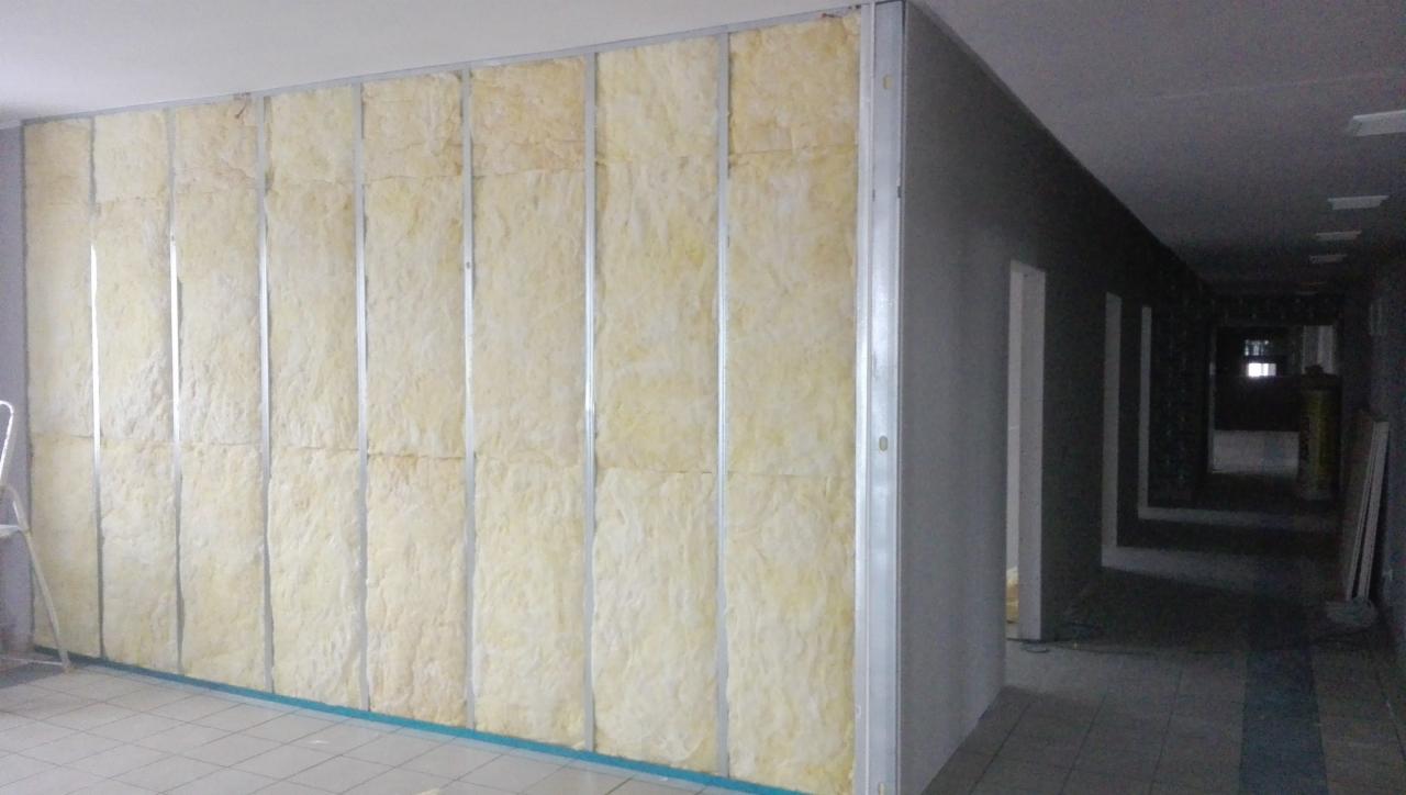 Montaż izolacji z wełny mineralnej w metalowym stelażu na ścianie w korytarzu biurowym, widoczne elementy konstrukcyjne i fragment drabiny.