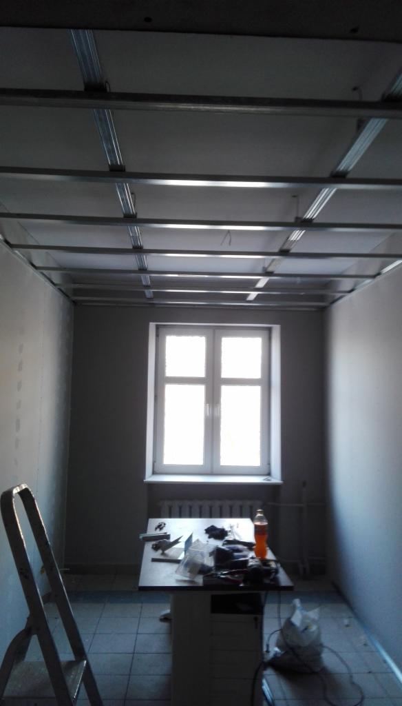 Montaż metalowego stelaża podwieszanego sufitu w pokoju z oknem, widoczne narzędzia na stole i drabina oparta o ścianę.