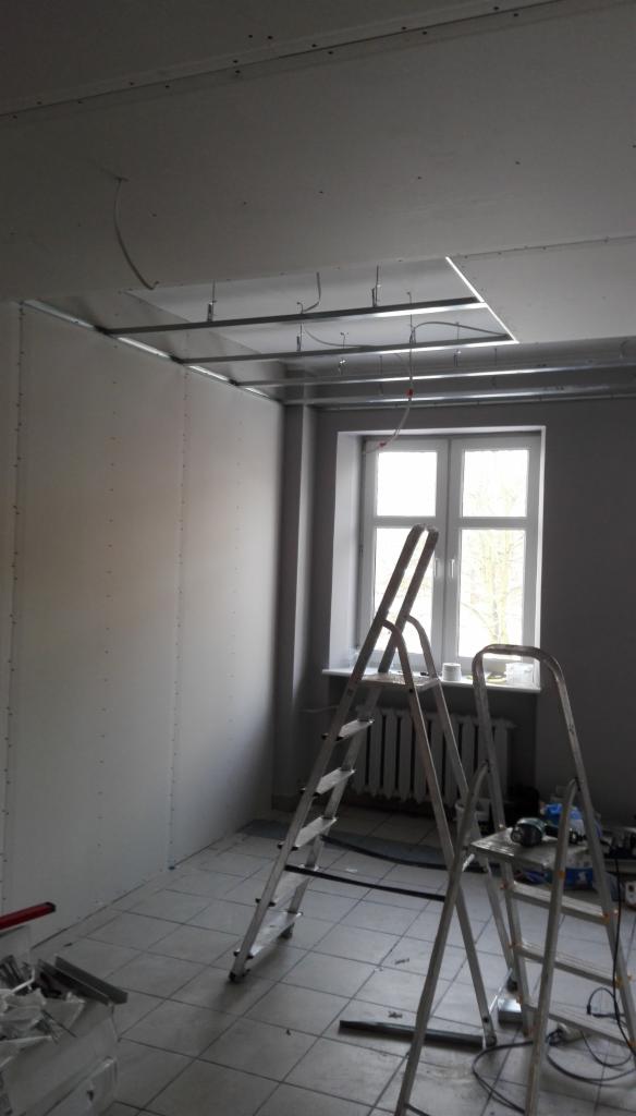 Wnętrze pokoju w trakcie montażu sufitu podwieszanego z płyt gipsowo-kartonowych, widoczne metalowe profile, częściowo zamontowane płyty oraz dwie aluminiowe drabiny.