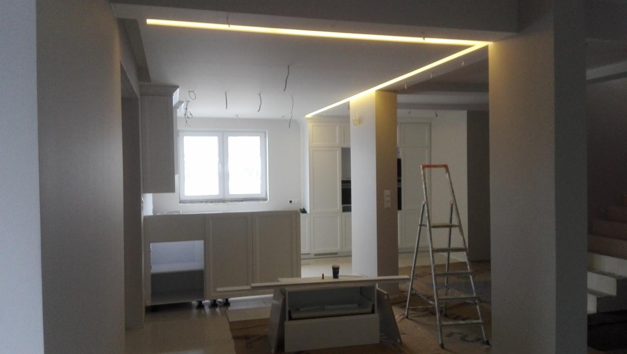 Wnętrze w trakcie prac wykończeniowych: zabudowa meblowa kuchenna w kolorze białym, podwieszany sufit z oświetleniem LED, widoczne kable i drabina, przygotowanie do malowania.