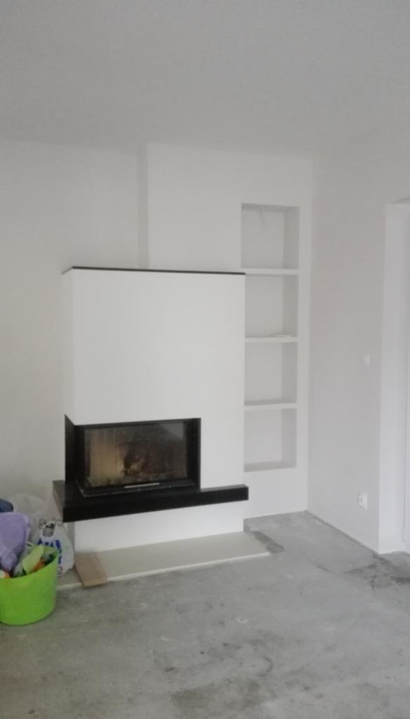 Nowoczesny kominek z czarnym obramowaniem w białej obudowie, w tle wnęki w ścianie, podłoga w trakcie prac wykończeniowych, widoczne wiadro z narzędziami malarskimi.