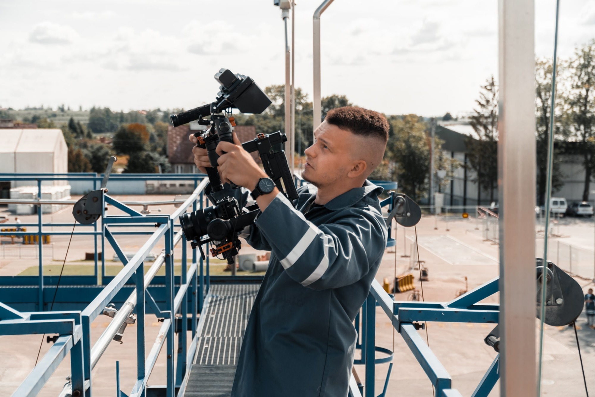Operator w kombinezonie, stojący na konstrukcji stalowej, obsługuje profesjonalny stabilizator kamery z zamontowanym monitorem, przygotowując się do nagrania ujęcia z wysokości.