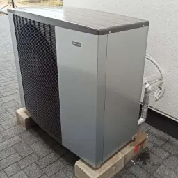 Szara pompa ciepła Viessmann stojąca na betonowych bloczkach na szarej kostce brukowej przy białej ścianie budynku.