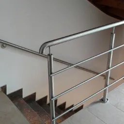 Wewnętrzna balustrada ze stali nierdzewnej z poziomymi prętami, zamontowana przy schodach z ciemnego kamienia, widoczna z góry, z drewnianymi schodami spiralnymi w tle.