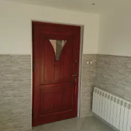 Wnętrze przedpokoju z wykończonymi ścianami w szare płytki imitujące kamień, ciemnoczerwone drzwi z szybką w kształcie trójkąta, biały grzejnik żeberkowy i szara podłoga z połyskiem.