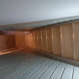 Wnętrze z zamontowanymi panelami podłogowymi w odcieniach szarości, widoczna drewniana konstrukcja antresoli z panelami na ścianach i suficie, oraz fragment buta roboczego w prawym górnym rogu.