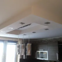 Wykończony sufit podwieszany z oświetleniem punktowym i chromowanymi lampami wiszącymi w nowoczesnej kuchni z oknem i zabudową meblową.