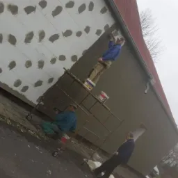 Pracownicy na rusztowaniu tynkują elewację budynku, widoczne ocieplenie z nałożonymi plackami kleju, jeden z pracowników stoi na rusztowaniu i nakłada tynk, drugi siedzi na rusztowaniu i również...