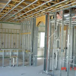 Wnętrze budynku w trakcie budowy, widoczne metalowe profile ścian działowych, instalacje elektryczne i sanitarne w surowym stanie, drewniana konstrukcja stropu podwieszana na metalowych profilach...