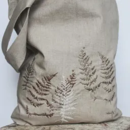Beżowa, lniana torba na ramię z brązowym i białym haftem przedstawiającym motyw paproci, ustawiona na postarzanym, drewnianym stołku.