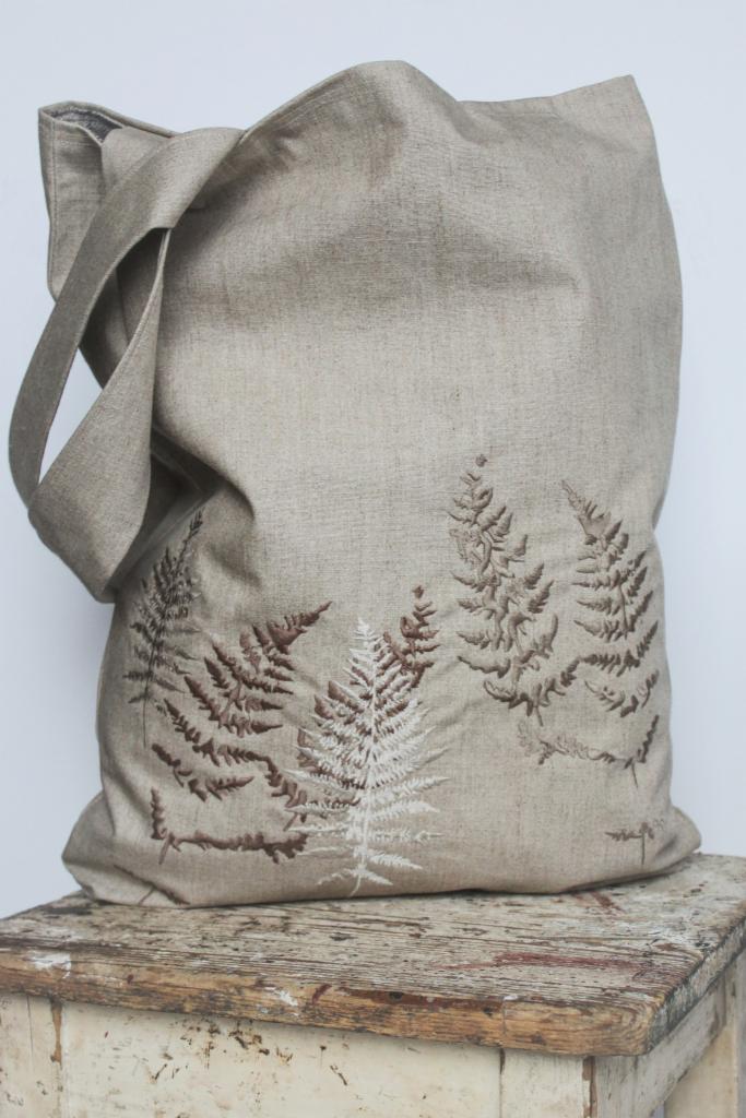 Beżowa, lniana torba na ramię z brązowym i białym haftem przedstawiającym motyw paproci, ustawiona na postarzanym, drewnianym stołku.