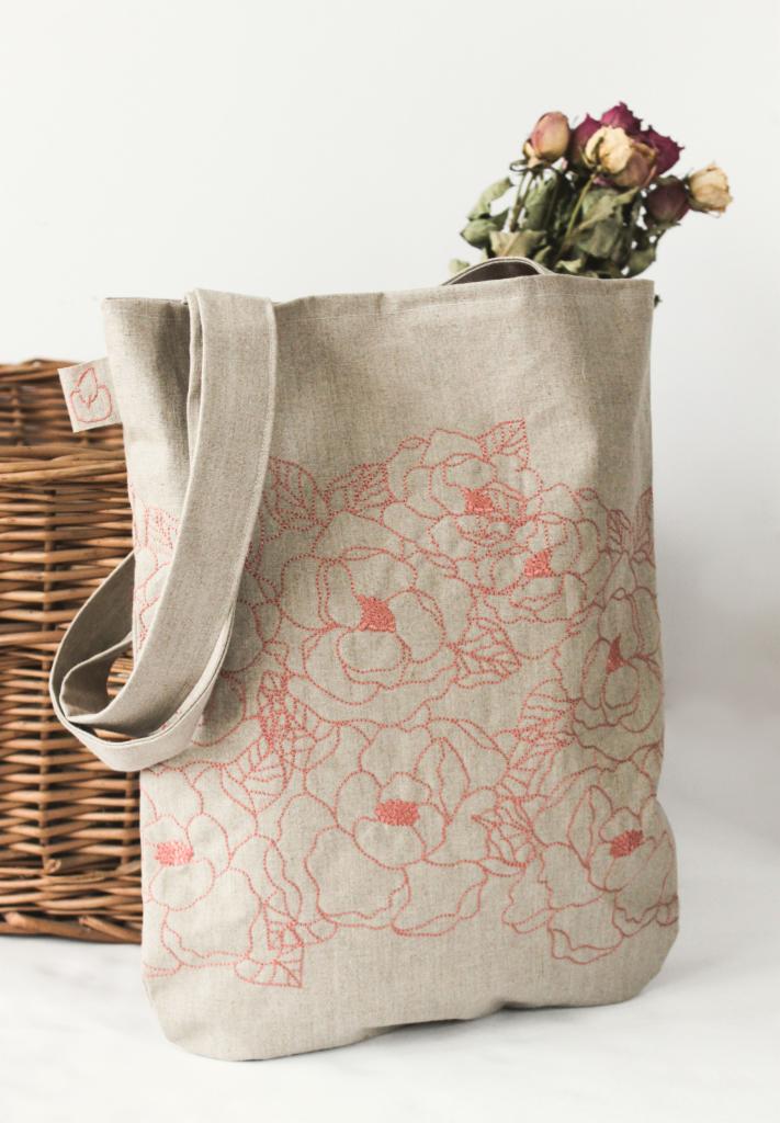 Lniana torba na zakupy z delikatnym haftem w kwiaty, umieszczona obok wiklinowego kosza i bukietu suszonych róż.