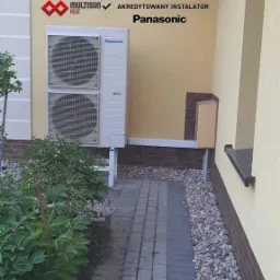 Zewnętrzna jednostka pompy ciepła Panasonic zamontowana na elewacji budynku, widoczne logo firmy instalacyjnej 'PRO Partners' i certyfikat 'Akredytowany Instalator'.