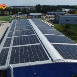 Duża instalacja paneli słonecznych na dachu hali przemysłowej, widok z góry, logo firmy w lewym górnym rogu.