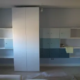 Biała zabudowa meblowa z szafą dwudrzwiową, otwartymi półkami i szafkami w odcieniach błękitu, na tle szarej ściany w pokoju.