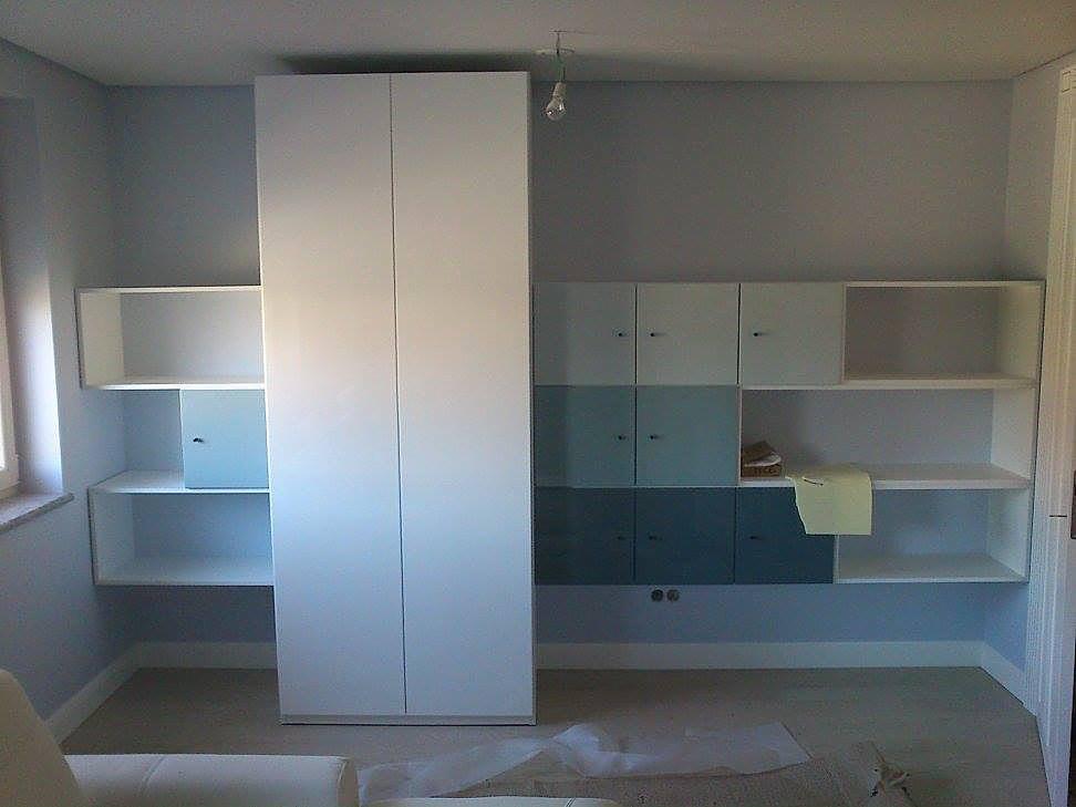 Biała zabudowa meblowa z szafą dwudrzwiową, otwartymi półkami i szafkami w odcieniach błękitu, na tle szarej ściany w pokoju.