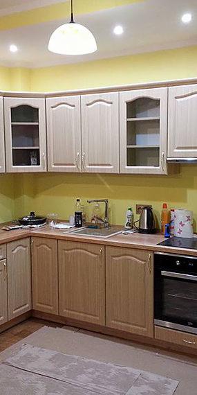 Jasna kuchnia z drewnianymi frontami szafek, górne szafki częściowo przeszklone, żółte ściany, na podłodze zabezpieczenie remontowe.