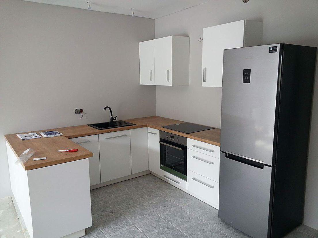 Wnętrze kuchni w trakcie montażu: białe szafki, drewniany blat, czarny zlew i srebrna lodówka, widoczne niedokończone wykończenia ścian i podłogi, narzędzia na blacie.