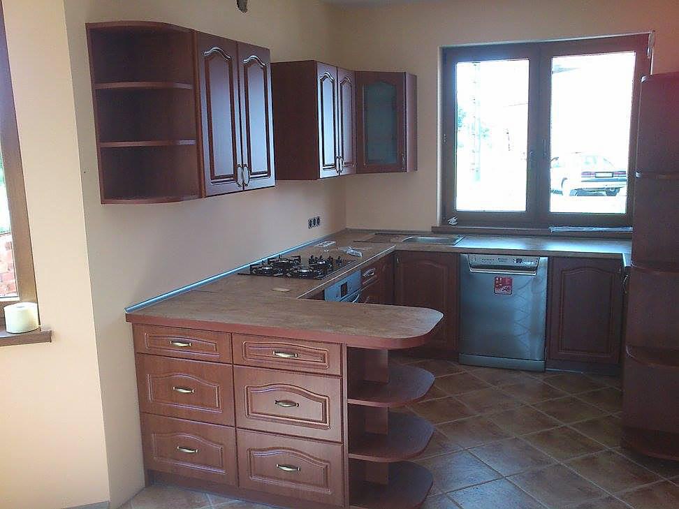 Klasyczna kuchnia z drewnianymi frontami w odcieniach brązu, widoczna płyta gazowa, zlew i zmywarka, na podłodze brązowe płytki, za oknem zaparkowany samochód.