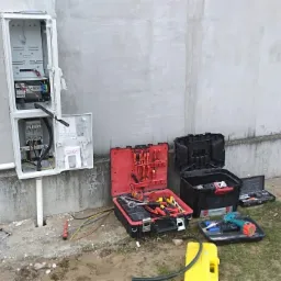 Otwarta skrzynka elektryczna na szarej ścianie budynku, z widocznymi przewodami i elementami instalacji, obok rozłożone skrzynki z narzędziami elektrycznymi i akcesoriami.