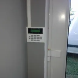 Panel sterowania alarmem domowym zamontowany na szarej ścianie, widoczny fragment otwartych białych drzwi i zaparkowany biały samochód dostawczy na zewnątrz.