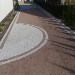 Ułożona z kostki brukowej ścieżka z wyznaczonymi pasami z jasnej kostki, obrzeża z roślinami i kamieniami wzdłuż ściany budynku.