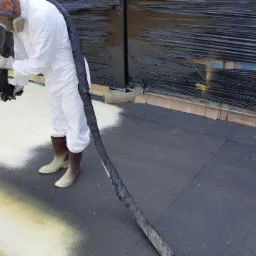 Pracownik w kombinezonie ochronnym aplikuje piankę PUR na dach w Lubinie, widoczny fragment dachu pokryty pianką w kolorze żółtym i białym, w tle zabezpieczona folią ściana.