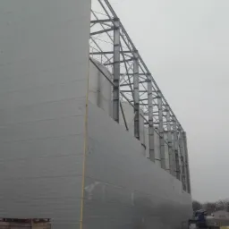 Montaż obudowy hali z płyt warstwowych i stalowej konstrukcji szkieletowej, widoczne silosy wewnątrz, podnośnik koszowy w tle.